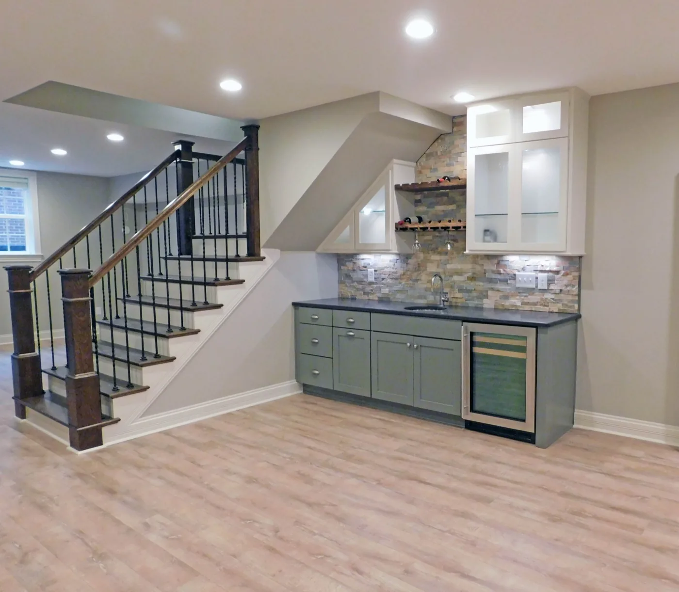 A finished basement with a wet bar, stone backsplash and a stained oak stair rail